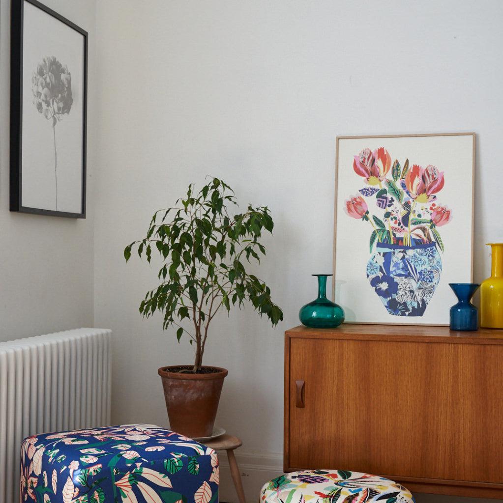 Stylish interior featuring a mid-century sideboard adorned with colourful vases and a framed Blue China Vase fine art print by Kitty McCall. The room is complemented by bold floral pouffes, patterned cushions, and a lush potted plant, creating a vibrant and inviting decor.