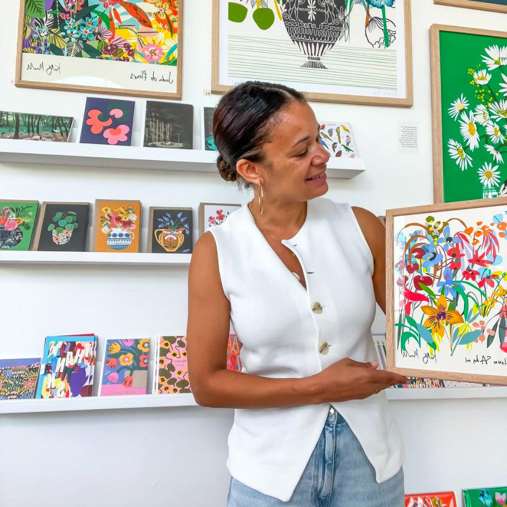 Woman holding an A4-sized Flower Study No. 1 fine art giclée print by Kitty McCall, surrounded by vibrant art prints and colourful cards displayed in a bright and modern gallery setting.