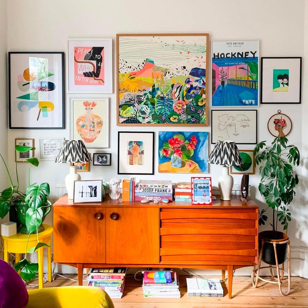 Bright and eclectic living space featuring a mid-century modern sideboard adorned with books, decorative lamps, and plants. A colourful gallery wall above showcases vibrant art prints, including floral, abstract, and landscape designs, creating a lively and stylish interior