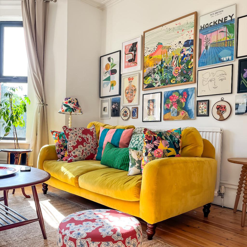 Bright and colourful living room featuring a yellow velvet sofa adorned with vibrant patterned cushions, a red floral ottoman, and a gallery wall of eclectic art prints, creating a cheerful and stylish interior design.