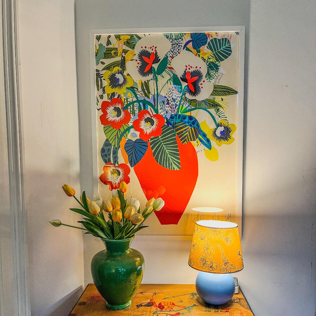 Red Vase fine art print by Kitty McCall displayed on a wall, featuring bold red and vibrant floral patterns in a modern setting, complemented by a green vase of tulips and a decorative yellow lamp on a vintage wooden chest